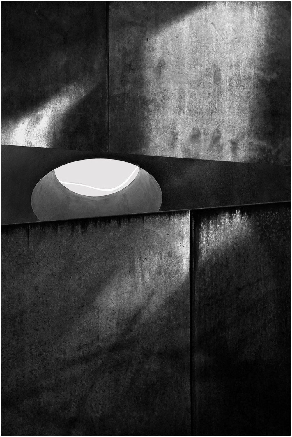 Bildmontage in Schwarz-Weiß: Eine Betonwand, in der sich im oberen Drittel ein dunkelgrauer Keil befindet, in dem ein Loch eingelassen ist mit einer bandförmigen Lichtquelle.