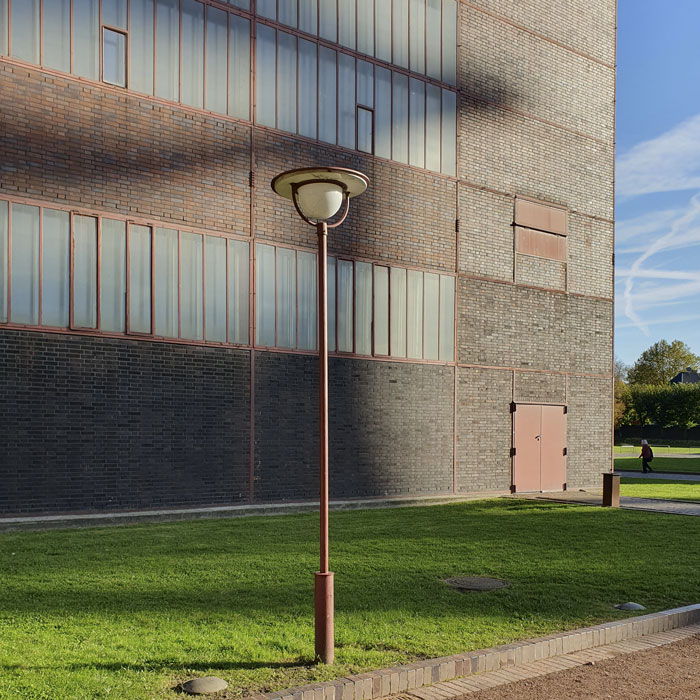 Farbfotografie, die eine Laterne vor einem Gebäude auf dem Terrain der Essener Zeche Zollverein im Sonnenlicht zeigt.
