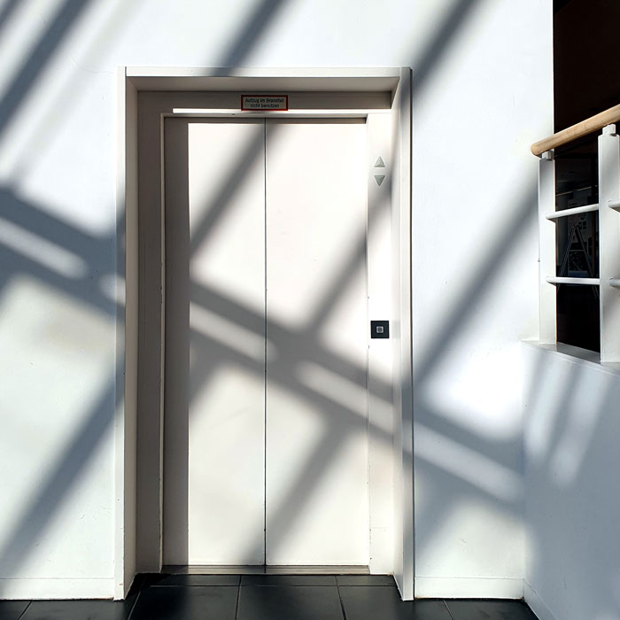 Farbfotografie, die eine Aufzugstür zeigt, in der die Umgebung Schatten in Form von schrägen Linien wirft.